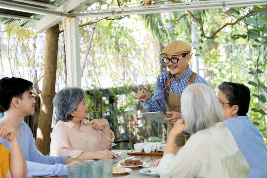 Senior friends and family pause at an outdoor cafe table while a smiling man takes their group photo, capturing reunion joy, shared mealtime and social connection across generations, aging society.