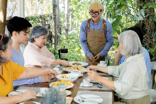 Senior friends receive food from a waiter at an outdoor cafe table while a young man helps nearby, creating a candid scene of service, conversation and multigenerational reunion dining, aging society.