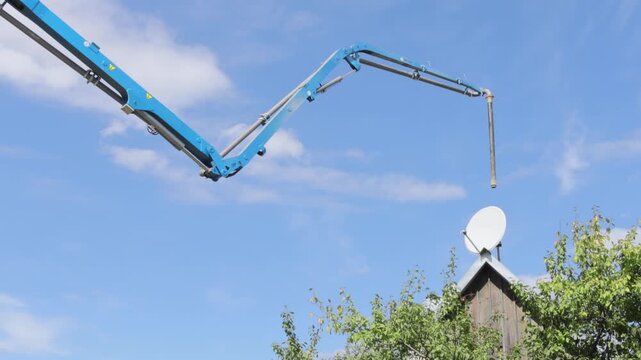 Blue concrete pump arm stretches across sky above rooftop. Jointed boom and dangling hose move over trees and satellite dish, showing active pumping setup on bright day with scattered clouds.