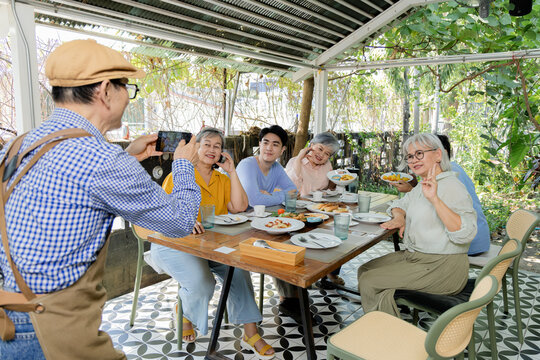 A cafe waiter leans across the outdoor table to serve bread and snacks to senior friends and family, capturing attentive service, shared food and lively reunion interaction outdoors, aging society.