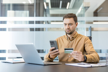 Obraz premium Serious man using a smartphone and holding a payment card, sitting at a desk with a laptop, engaging in secure mobile banking or e-commerce transaction in a modern office environment