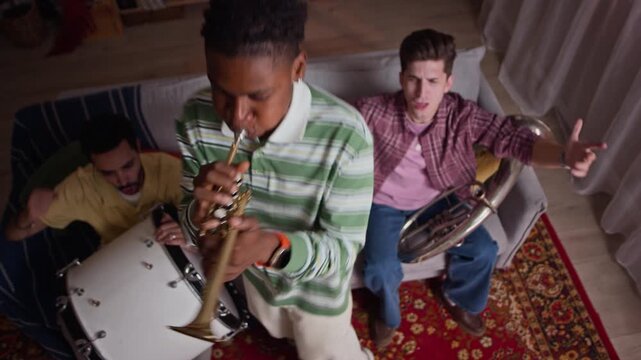 Diverse young adult male football fans playing trumpet and drums at home in slow motion, high angle shot. Visual asset perfect for sports fandom campaigns and youth culture commercials