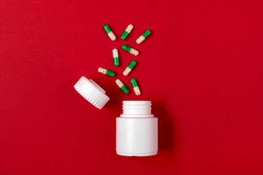 White plastic pill bottle with scattered green and white capsules on a vibrant red background, ideal for healthcare and pharmaceutical concepts.