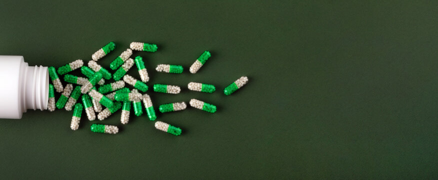 White and green medicine capsules spill from a bottle onto a textured green surface, suitable for healthcare, pharmacy, and medical concepts.