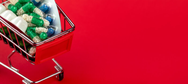 Mini shopping cart filled with various medication capsules and blister packs on vibrant red background, symbolizing pharmaceutical retail and health care choices.