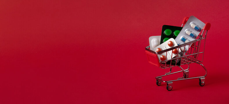 Mini shopping cart filled with blister packs of pills on a vibrant red background, symbolizing healthcare, pharmacy, and pharmaceutical shopping.