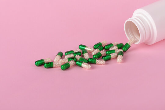 Green and white capsules spilling from a white bottle onto a pastel pink surface, ideal for healthcare and pharmaceutical themes.
