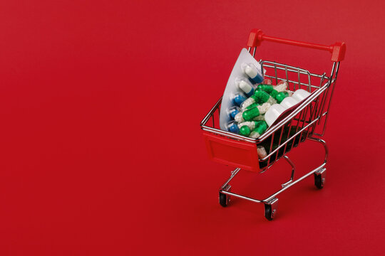 Miniature shopping cart filled with assorted pills and capsules on red background, symbolizing healthcare, pharmacy, and medication shopping.