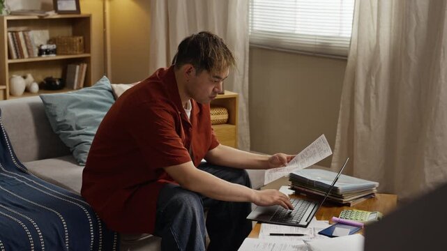 Asian young adult man sitting on sofa, holding utility bills and typing on laptop to pay expenses online, zoom in. Clip is perfect for banking apps, financial planning, or debt management ads