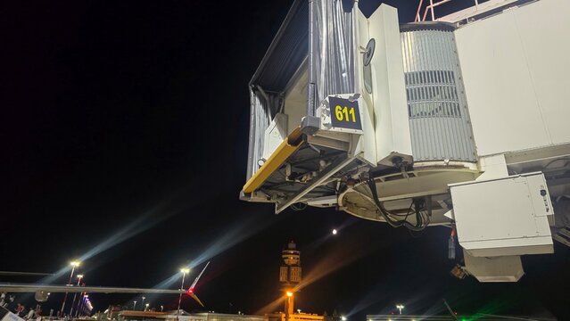 Airport jet bridge and boarding gate stand illuminated at night beside an aircraft. Concepts: air travel, aviation infrastructure, boarding, transportation, tourism, nighttime operations.
