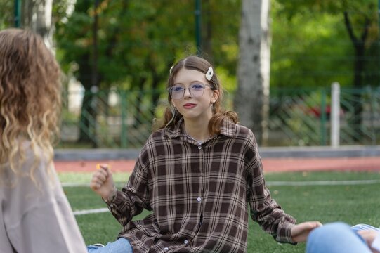 Teenage high school girl with curly hair talks with a classmate while sitting on an outdoor school sports field. 