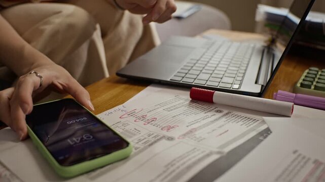 Young adult Caucasian woman typing on laptop, checking smartphone and highlighting utility bills with red marker. Clip is ideal for commercial videos about personal finance and home budgeting