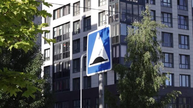 Blue-and-white road sign indicating artificial speed bump stands in foreground. Modern apartment building and leafy trees form urban backdrop