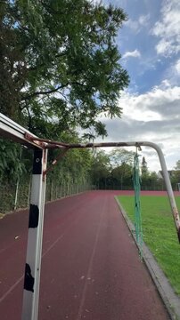Low angle view along a rusty white goal frame with a torn green net above an empty red running track, vertical video of a weathered school sports ground and neglected public infrastructure
