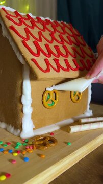 Vertical 4K video. 60 fps. Female hand decorates gingerbread house with wavy pattern on cookie roof, white edible paint apply icing to window pretzel. Plastic bag. Sugar food decor for Happy New Year