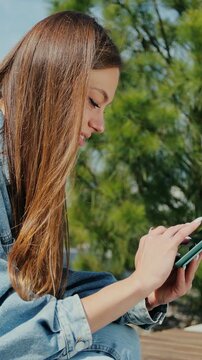 Vertical video. Woman is sitting on a park bench and scrolling on the tablet in her hands. female messaging and chatting on tablet computer in city.