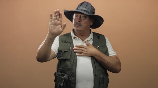 Man wearing fisherman's hat and fishing vest raises right hand palm forward and presses left hand to chest in studio setting facing camera; sincerity.