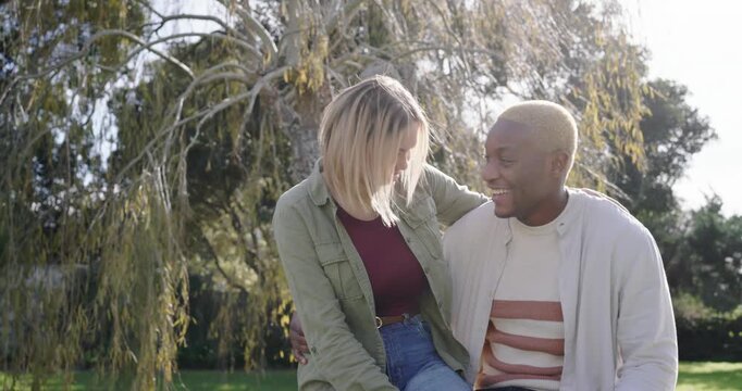 African American couple exchanging smiles sitting on park bench under weeping tree, woman rising