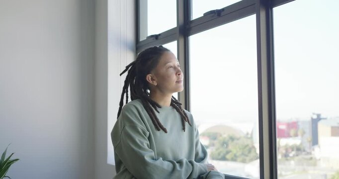 Non-binary person in green sweatshirt leaning by large window, responding to camera gaze, smiling