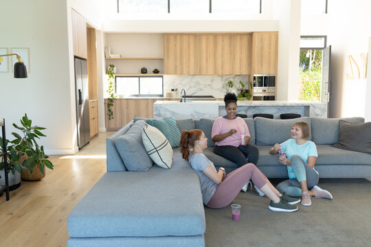 Diverse female friends chatting in sneakers on gray sofa in open plan living room with smoothies