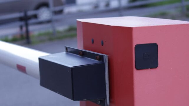 Close-up of a red automatic barrier post with white-and-red striped arm and black sensor module; blurred parked cars and building form background