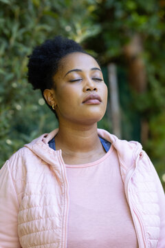 Adult woman posing in garden, closing eyes, tilting head, wearing pink vest, gold hoops, dark strap