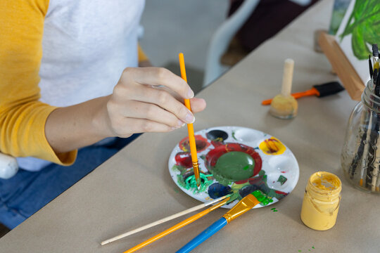 Female in white yellow-sleeved top mixing green paint with brush on palette at craft table