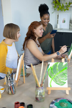 Three female friends painting at craft table in studio with easels, brushes, palette, smartphone