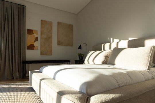 Large padded bed is sitting in sunlit bedroom while light is casting linear shadows across bedding