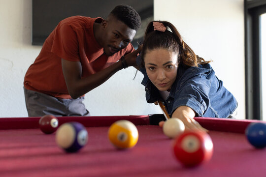 Diverse friends leaning over red-felt pool table at home, woman taking shot with cue, colored balls