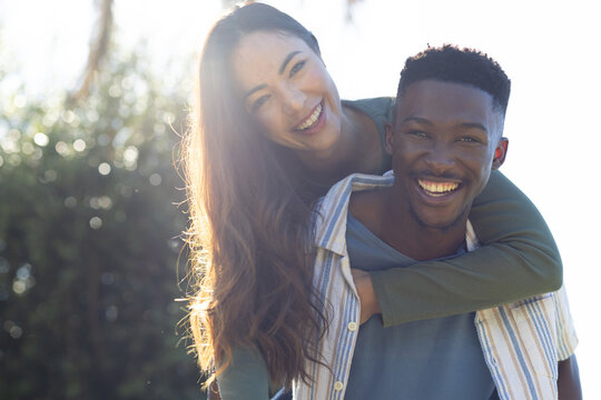 Diverse couple sharing piggyback ride in sunlit park, man wearing striped shirt, woman green top