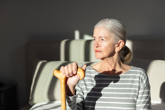Senior woman sitting on sofa at home wearing striped top holding cane facing right in sunlight