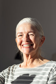 Senior woman smiling, wearing striped top and stud earrings, facing sunlight against gray wall