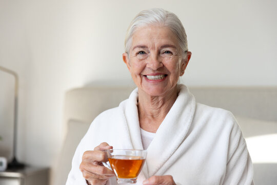 Senior woman smiling while holding clear glass teacup and wearing white bathrobe in bedroom