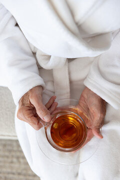 Senior woman holding glass teacup and saucer while sitting in white bathrobe on sofa