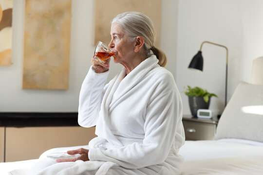 Senior female sitting on bed in white bathrobe holding clear glass cup and saucer sipping tea