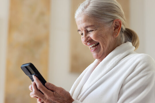 Senior woman in white robe smiling and laughing while holding clamshell mobile phone, copy space