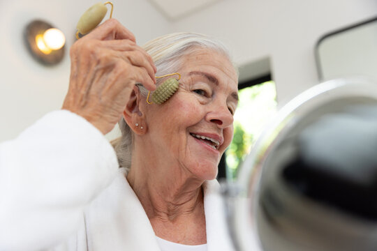 Senior woman sitting at vanity in white bathrobe using olive green facial roller near mirror