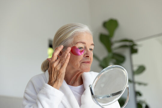 Senior woman wearing white robe applying pink hydrogel eye patch while holding round mirror at home