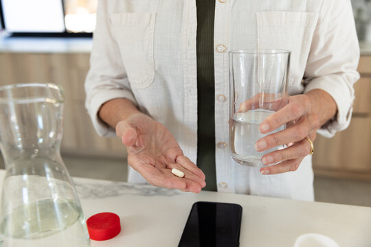 Senior woman wearing beige shirt over olive holding pill and glass at kitchen counter