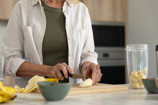 Senior woman trimming banana on cutting board and filling blender cup in kitchen wearing olive top