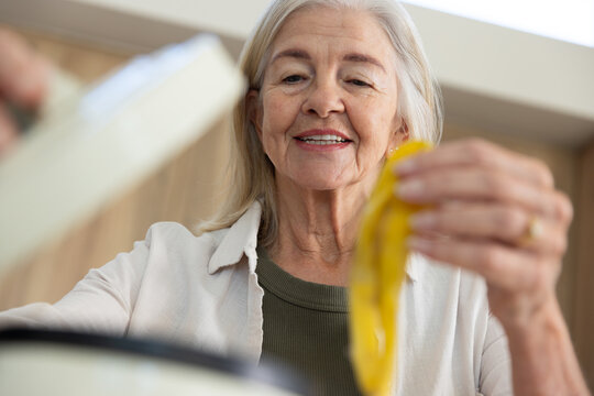 Senior female standing at kitchen counter holding banana peel and lid wearing button-up and ring