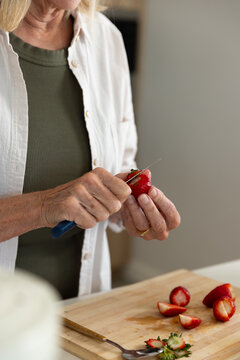 Senior woman holding, slicing strawberry with blue paring knife on cutting board at kitchen counter
