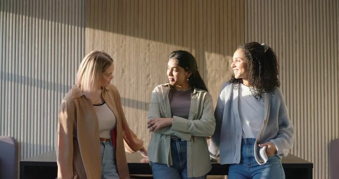Three women conversing by wood-slat wall with sunlit shadows, moving closer and posing for camera