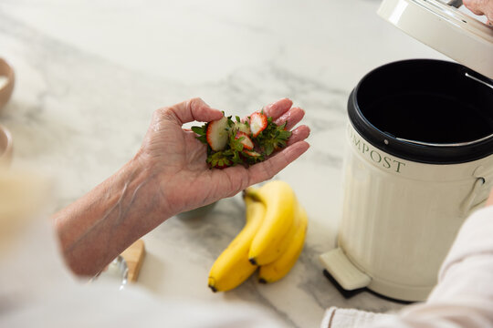 Senior woman holding strawberry tops and stems over compost bin on marble counter in light sweater