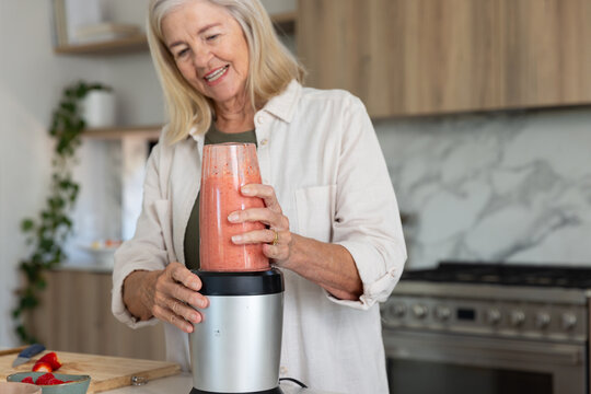Senior woman assembling pink smoothie at home kitchen counter with blender, cutting board
