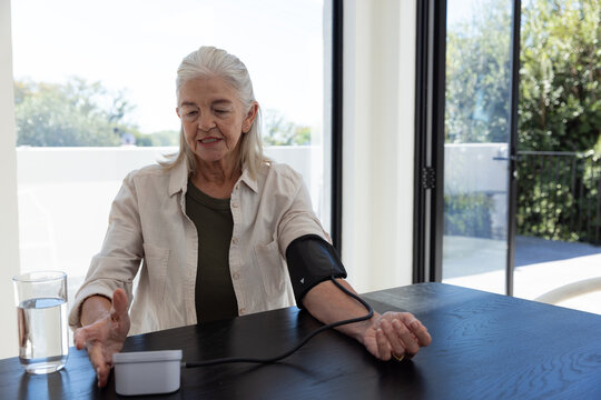 Senior woman sitting at dining-nook by doors, in beige shirt, checking pressure with cuff and meter