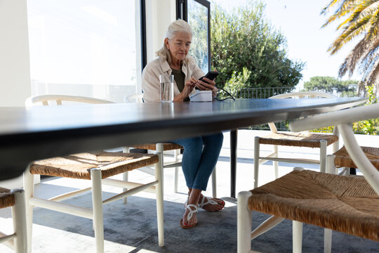 Senior woman sitting at dining table, wearing beige jacket and jeans, charging smartphone on dock