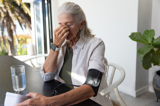 Senior woman sitting at home dining table wearing upper-arm blood pressure cuff holding monitor