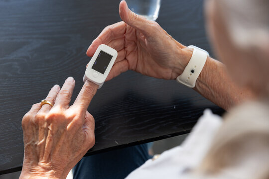 Senior woman holding pulse oximeter on left finger at dark wood table, wristband, ring, glass